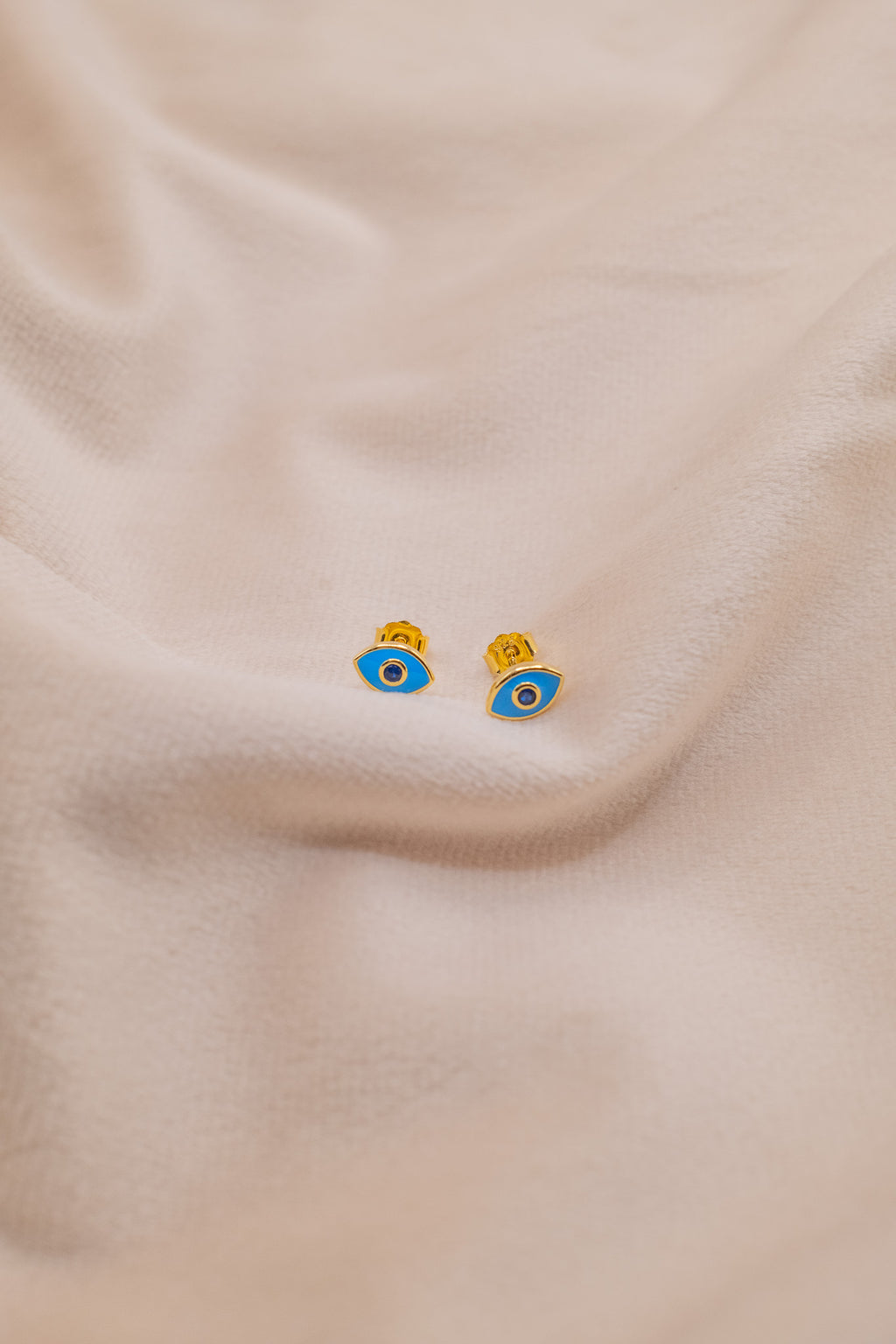 Zamari gold and blue eye-shaped earrings from an online dainty jewelry boutique on beige fabric